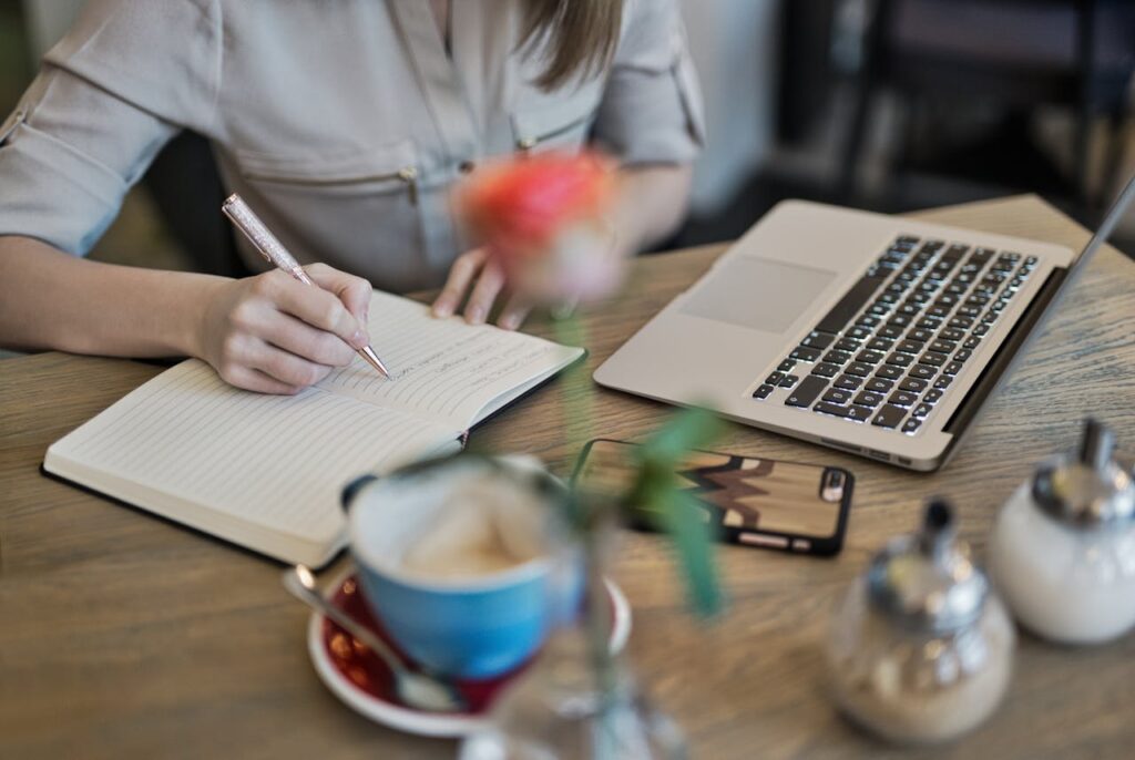 pexels photo 1766604 Woman writing in a notebook with a laptop and coffee cup on a desk. Ideal for workspace inspiration.